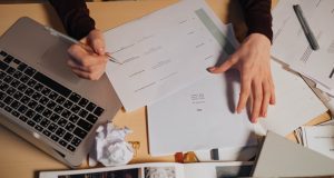 A person taking notes and working on a laptop surrounded by documents on a desk.